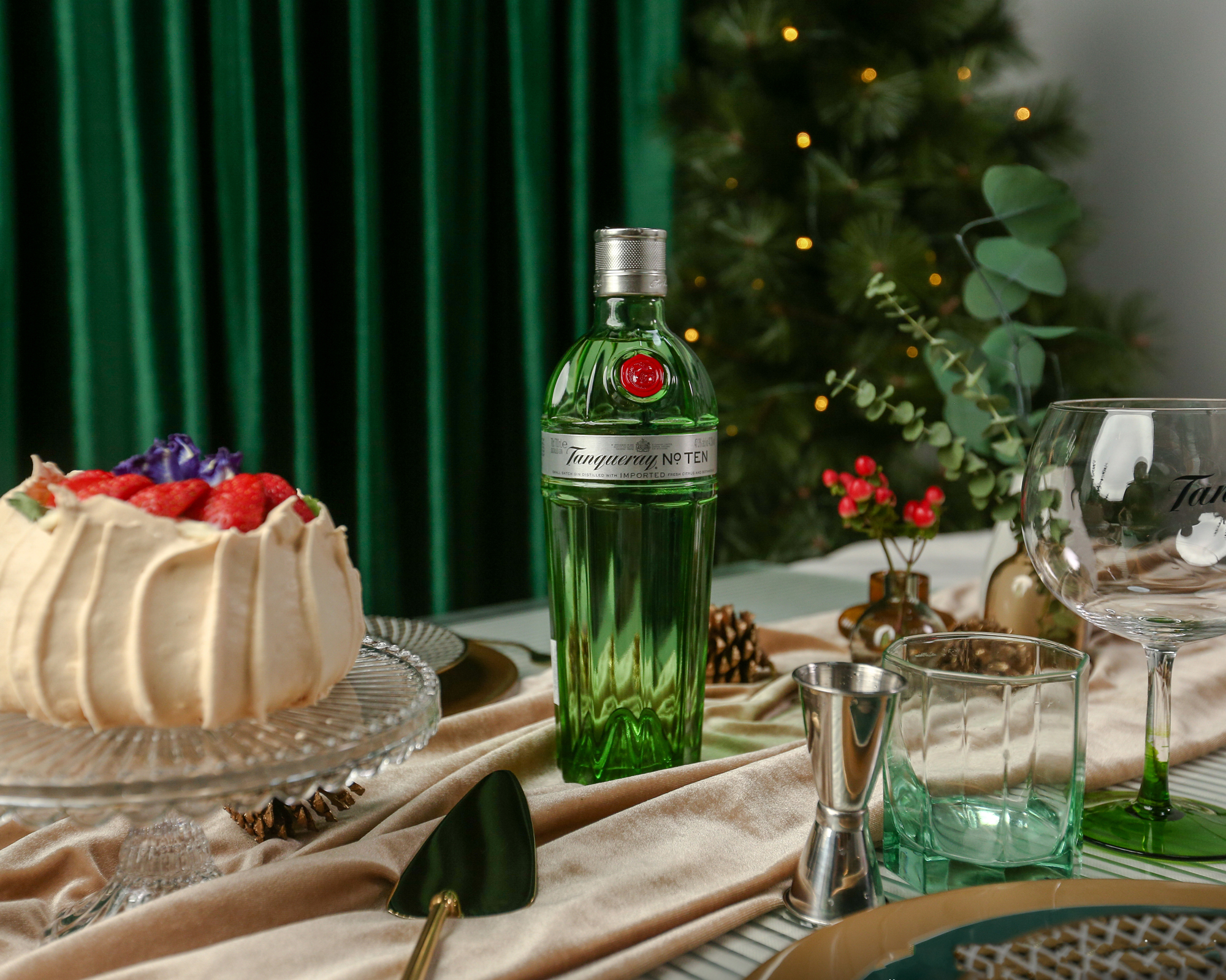 A bottle of Tanqueray No. Ten gin sits on a decorated table with a cake, glassware, utensils, and festive decor. A Christmas tree is visible in the background.