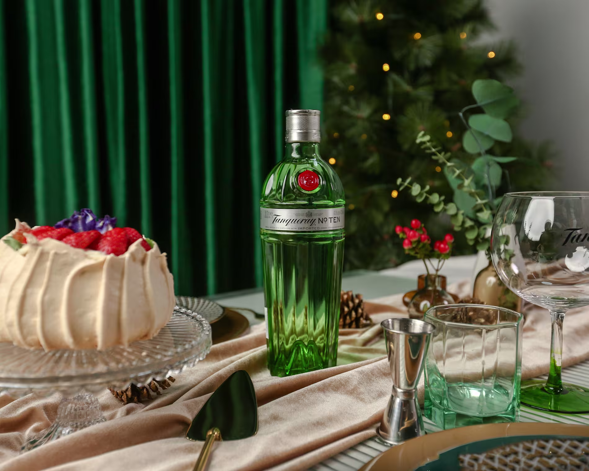 A bottle of Tanqueray No. Ten gin sits on a decorated table with a cake, glassware, utensils, and festive decor. A Christmas tree is visible in the background.