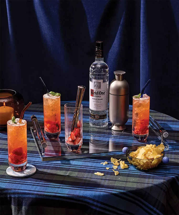 A bottle of Ketel One vodka, a cocktail shaker, three vodka cocktails with fruit, a bowl of chips, a candle, and utensils on a blue plaid tablecloth.