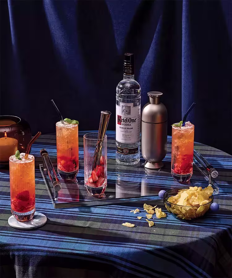 A bottle of Ketel One vodka, a cocktail shaker, three vodka cocktails with fruit, a bowl of chips, a candle, and utensils on a blue plaid tablecloth.