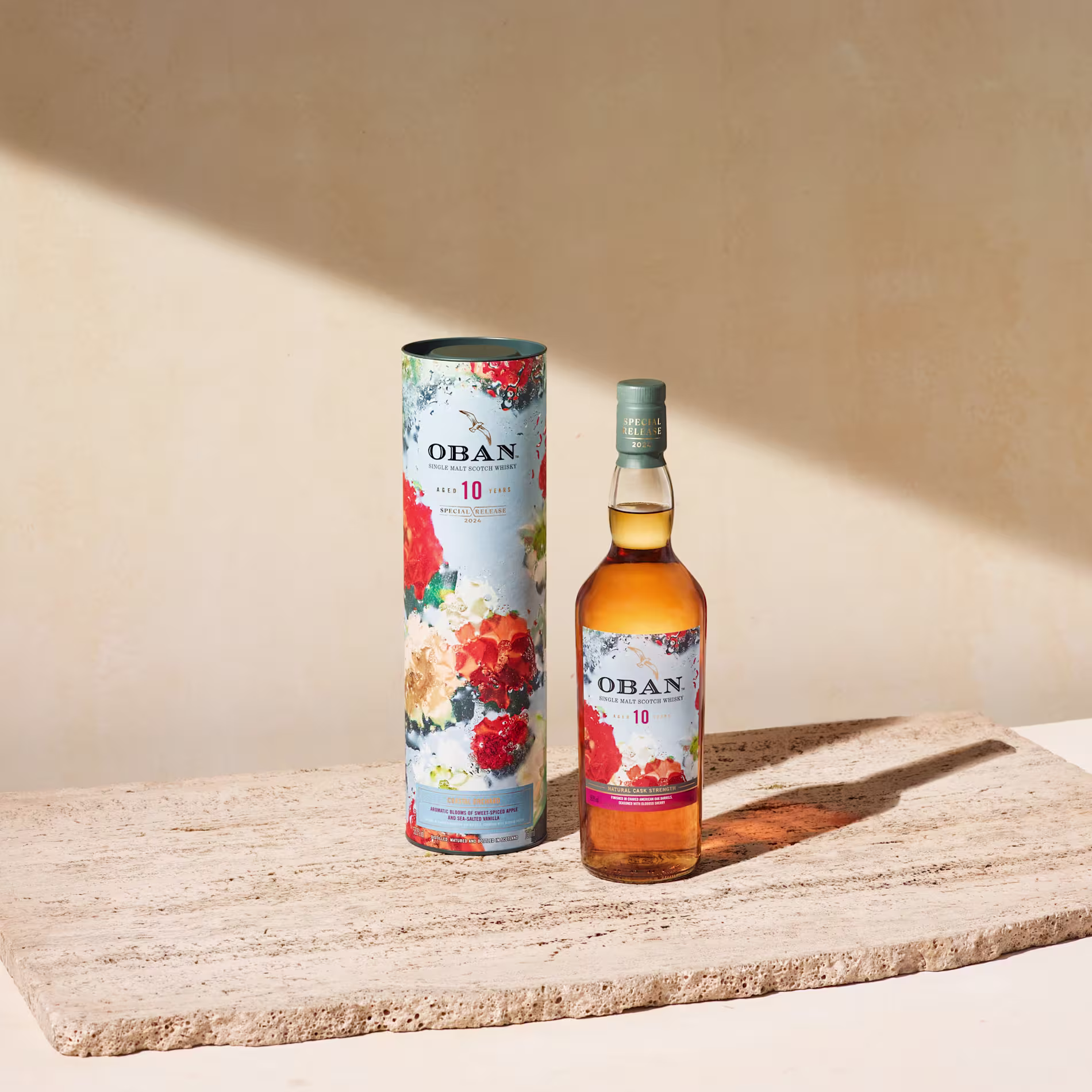 A bottle of Oban 10 Year Old whisky stands next to its colorful floral-patterned cylindrical box on a stone surface against a beige background.
