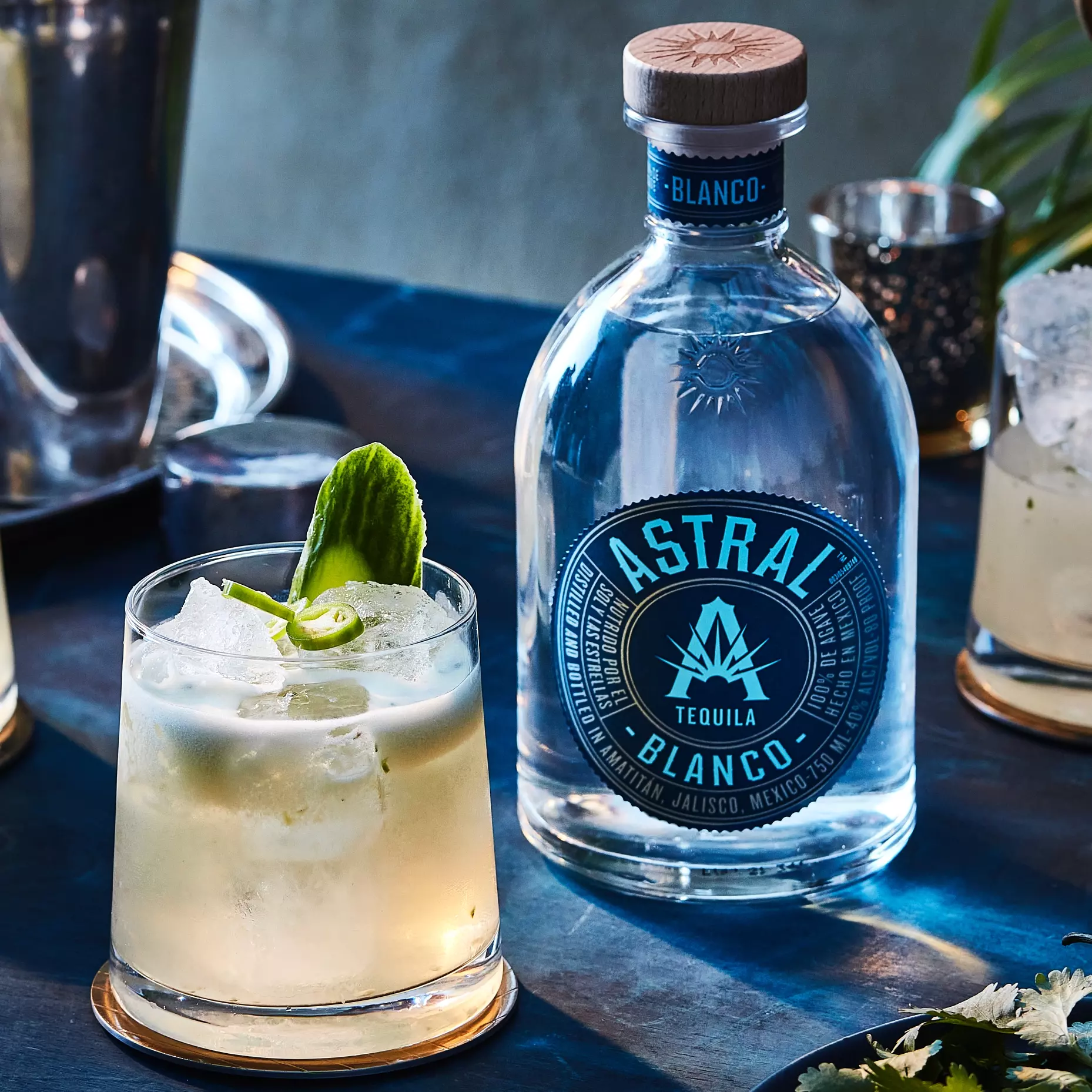 A bottle of Astral Blanco Tequila is next to a cocktail with ice, a cucumber slice, and a green garnish on a dark blue table.