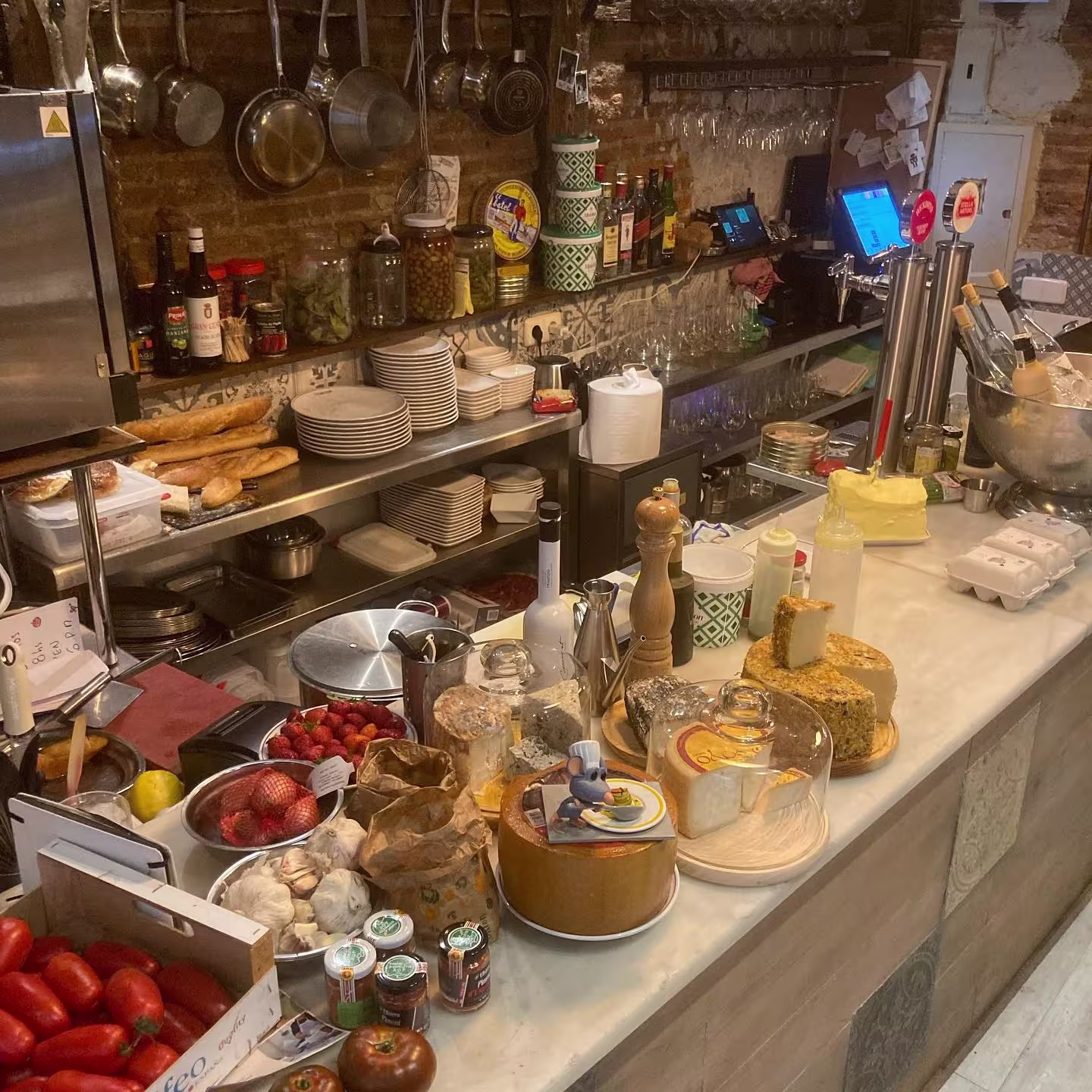La imagen muestra un mostrador de cocina de La Carbonería. Hay varios alimentos y utensilios de cocina visibles, incluyendo tomates, limones, frascos de condimentos, ollas y sartenes colgando, una tabla de cortar con pan y botellas de salsas.