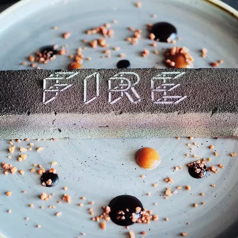 Fire - W Hotel, postre en forma de barra negra texturizada con el nombre "Fire", rodeado de salsas y crujientes que decoran un plato elegante.
