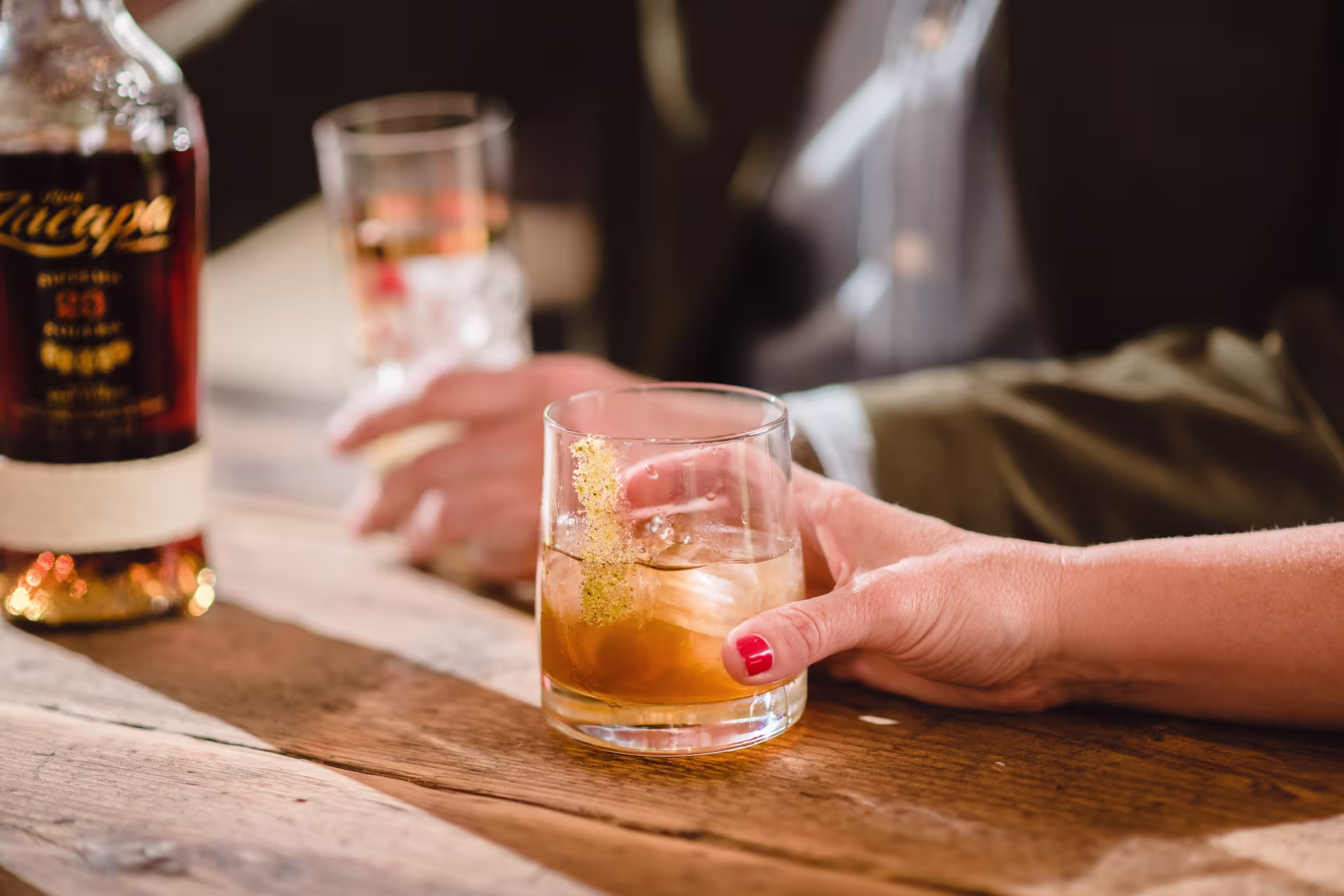 Close-up of a hand holding a glass of Ron Zacapa rum on the rocks, with a bottle and blurred background in a relaxed social setting