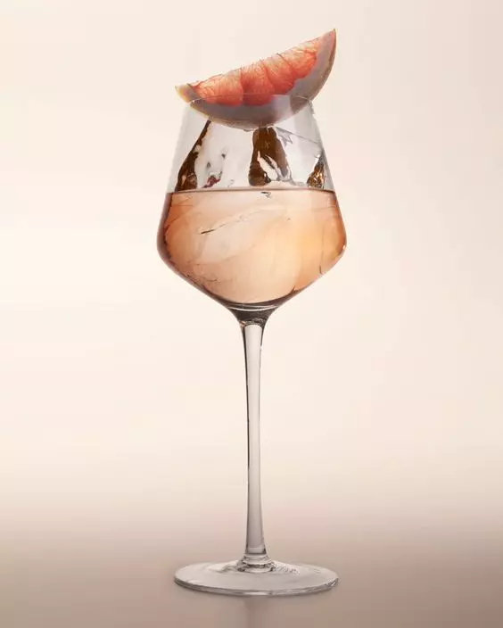 A stemmed wine glass filled with a light pink drink containing ice cubes, garnished with a wedge of grapefruit on the rim.
