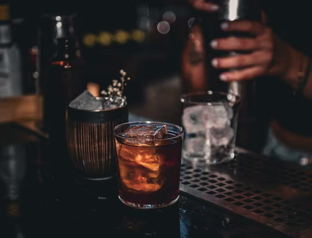 La imagen muestra un cóctel en un vaso corto con hielo, acompañado de otro vaso con hielo y una botella en la parte superior, mientras un bartender agita una coctelera en segundo plano.