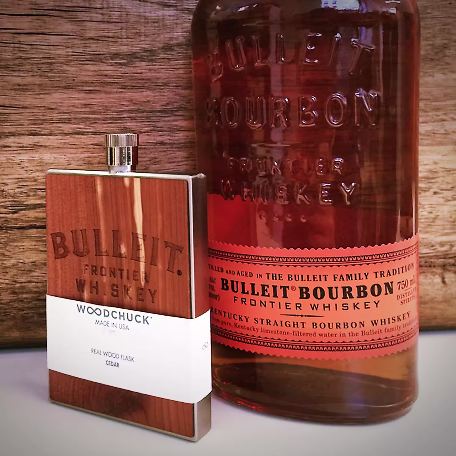 A bottle of Bulleit Bourbon whiskey stands next to a Woodchuck cedar wood flask labeled Bulleit Frontier Whiskey.