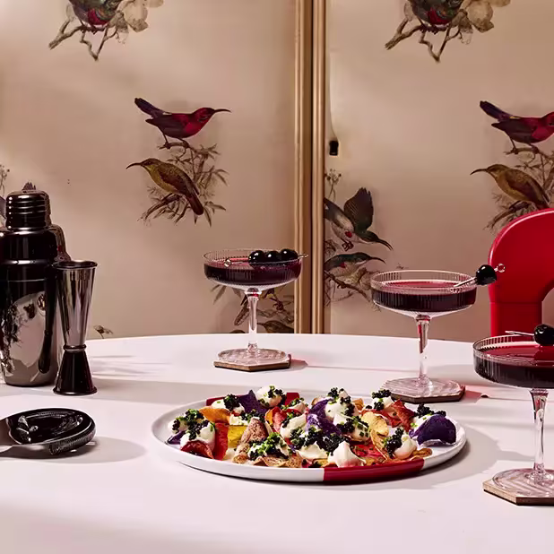 A table set with two cocktails garnished with cherries, a plate of assorted appetizers, a cocktail shaker, and a jigger, with a bird-patterned screen in the background.