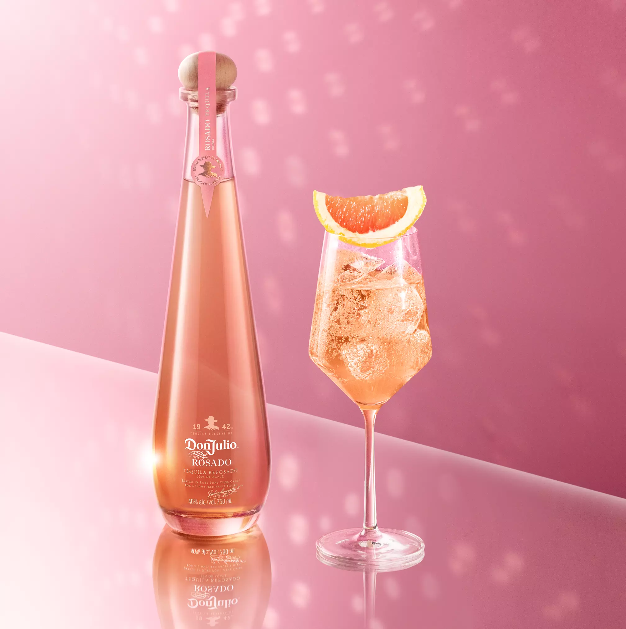 A bottle of Don Julio Rosado tequila stands next to a wine glass filled with an iced cocktail and a grapefruit wedge, set against a pink background with light reflections.