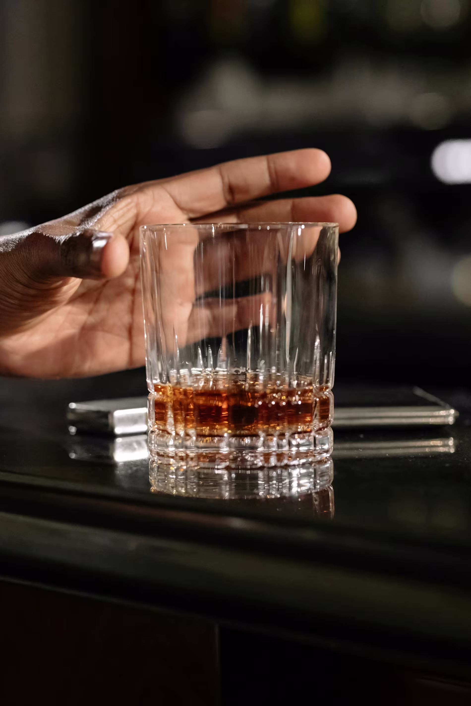 A hand reaches for a glass of brown liquor on a bar counter with a smartphone and another object in the background.