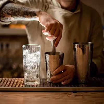 A bartender uses a muddler in a metal shaker while a glass filled with ice sits nearby on a bar counter.