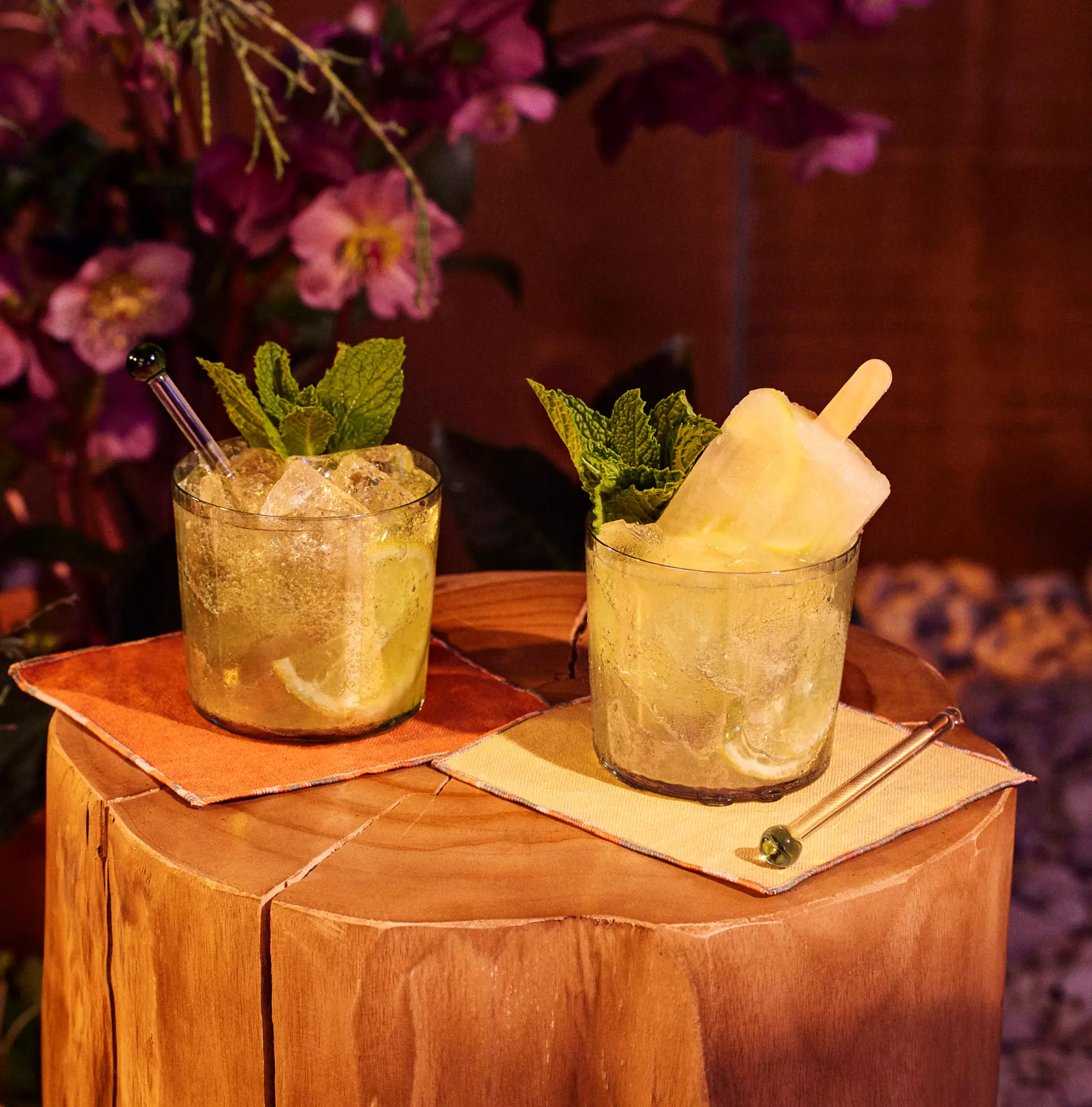 Two glasses of a mojito-style cocktail with lime, mint, and ice sit on a wooden surface; one glass contains a popsicle. Purple flowers are visible in the background.
