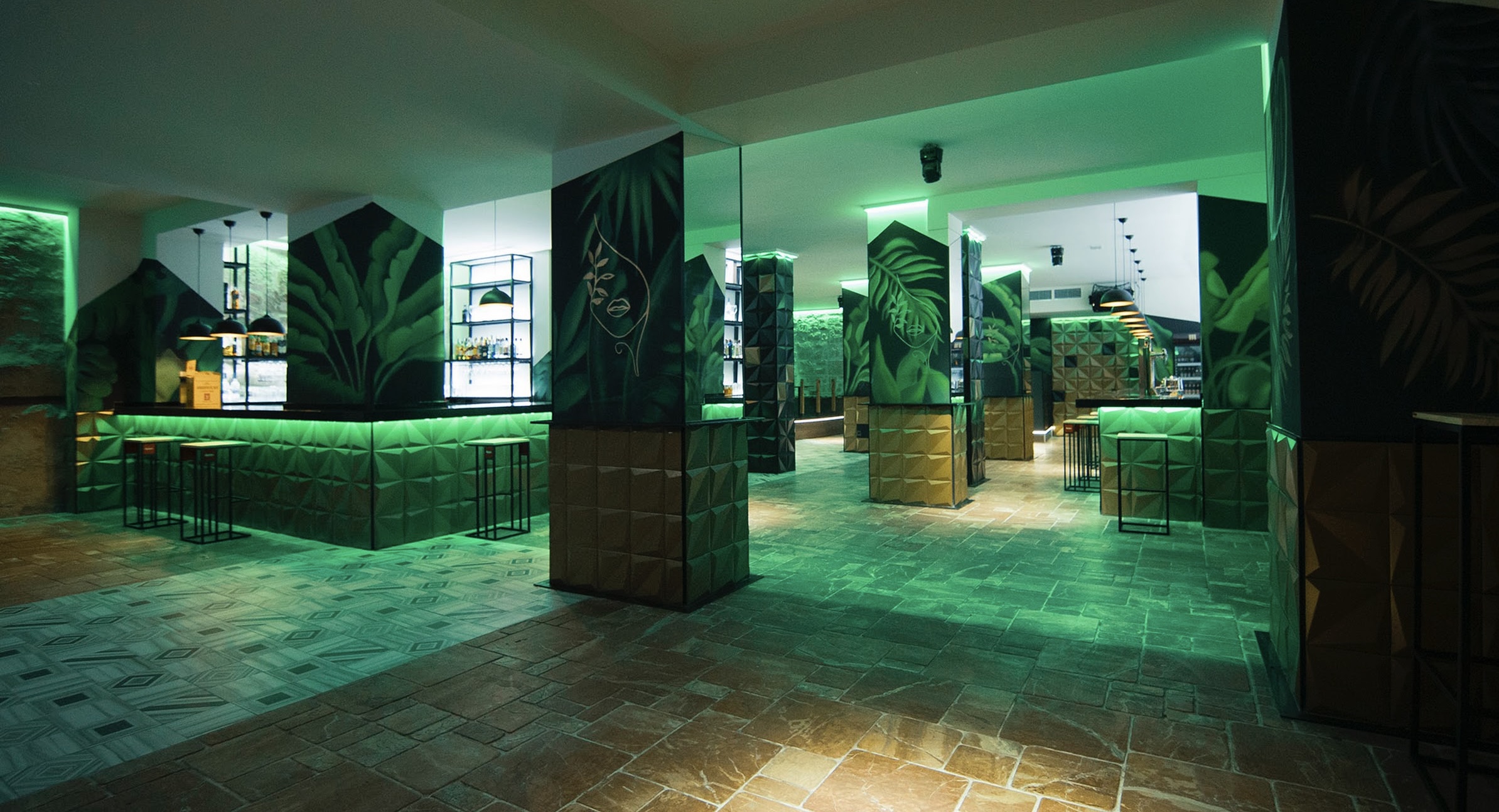 " La imagen muestra el interior de La Mona, un bar de estilo tropical y moderno. Con una iluminación verde envolvente, paredes decoradas con motivos de hojas y columnas geométricas, el espacio emana un ambiente fresco y exótico."