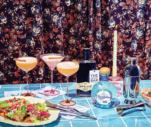 A tiled table with cocktails, a salad plate, cocktail shaker, bottles of Mr Black and Don Julio, a candle, bar tools, and floral curtains in the background.