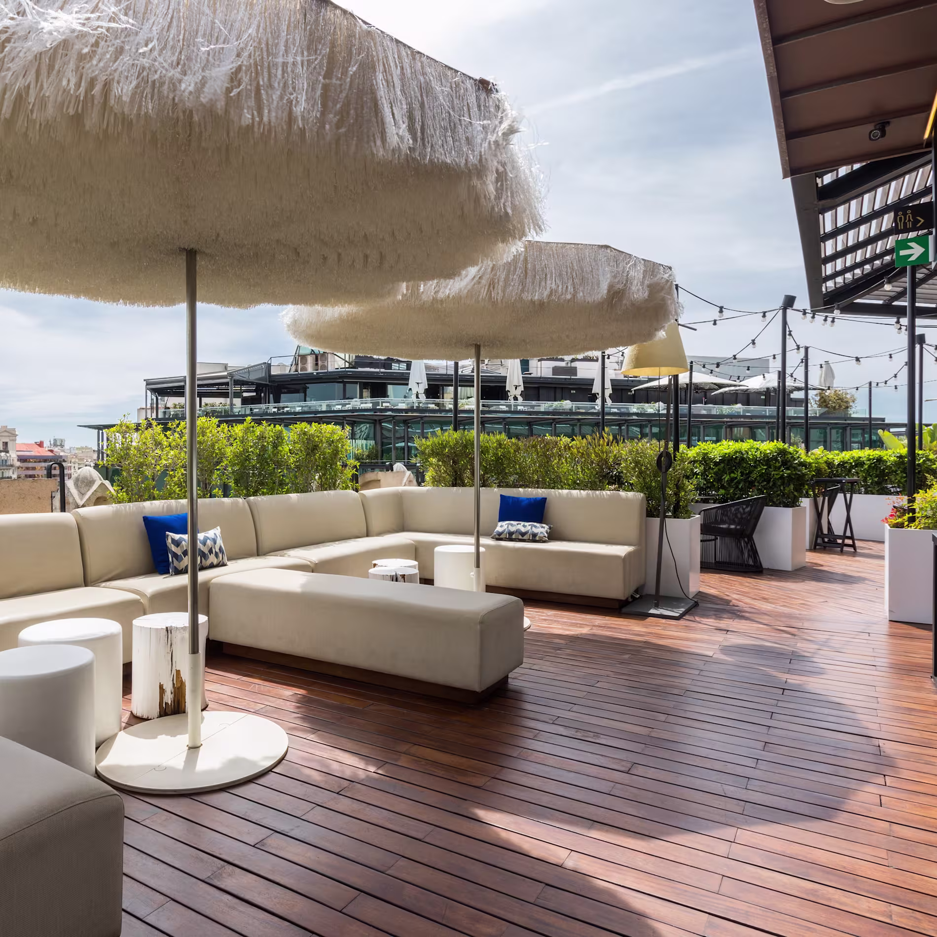La imagen muestra una terraza exterior de Verbena. Hay un gran sofá beige en forma de L con cojines blancos y azules, una pequeña mesa redonda blanca y taburetes cilíndricos blancos. Sobre la zona de asientos hay dos grandes sombrillas blancas