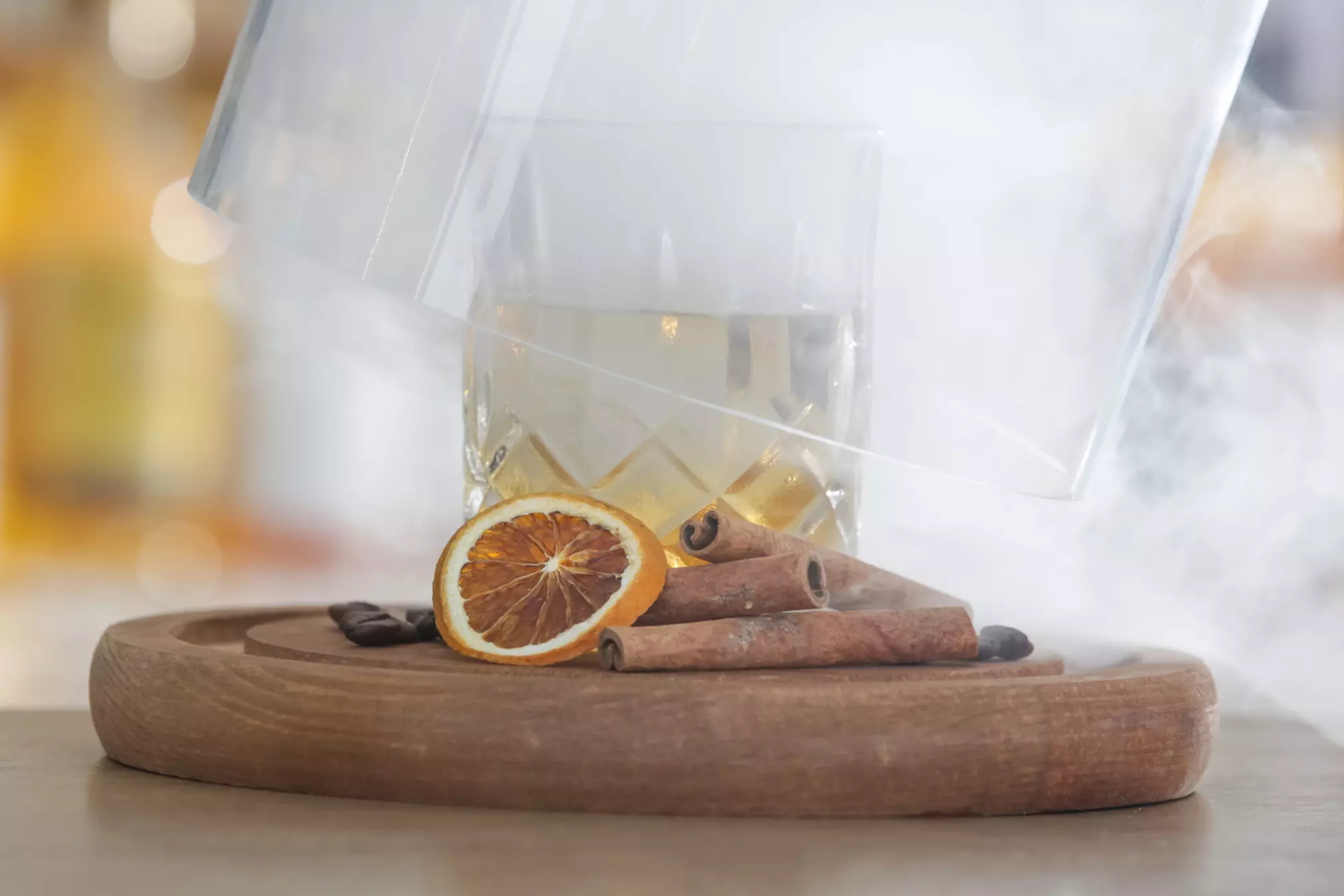 A glass of cocktail is partially covered by a glass dome with smoke, served on a wooden tray with dried orange slices and cinnamon sticks.