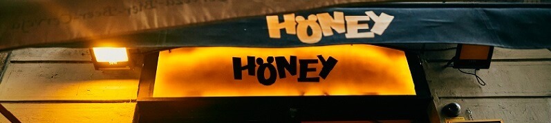 Fachada de Honey Furry Bar con su característico letrero amarillo iluminado y pizarras negras con ofertas de cócteles y cervezas artesanales.