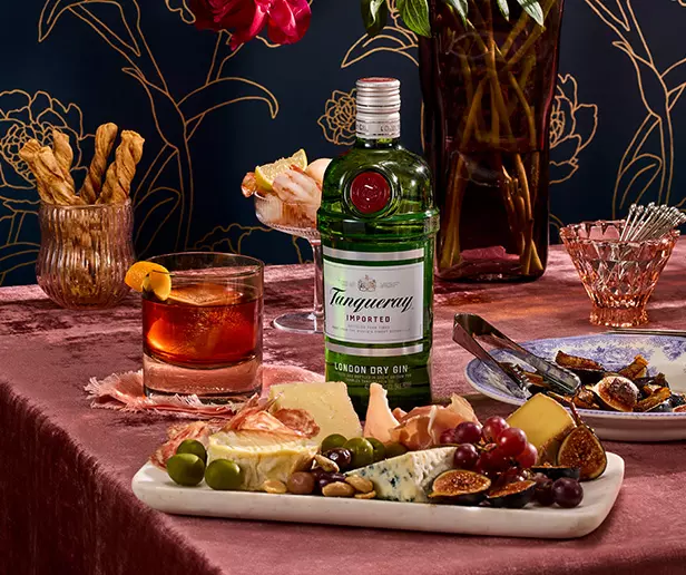 A bottle of Tanqueray gin, a cocktail, breadsticks, and a cheese platter with fruit and olives on a pink tablecloth.