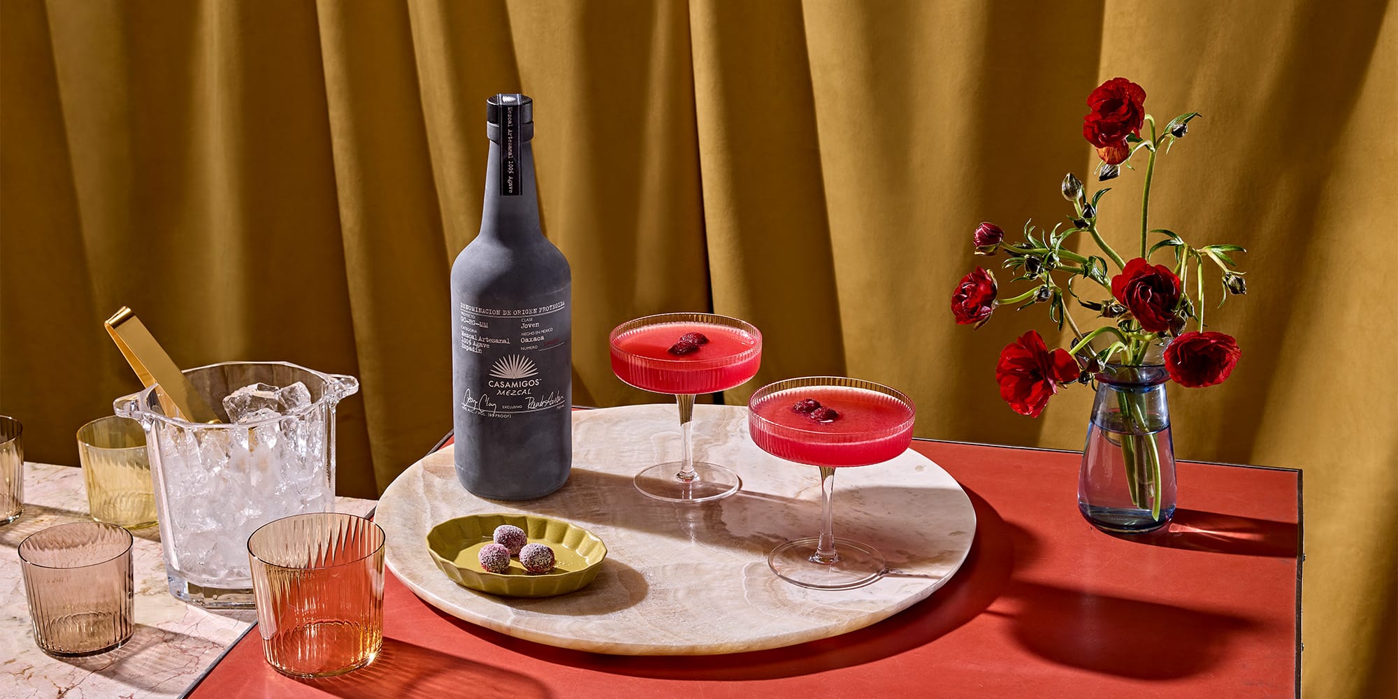 A marble tray with a dark bottle, two red cocktails in coupe glasses, a small plate with chocolates, and glassware, beside a vase with red flowers on a red surface.