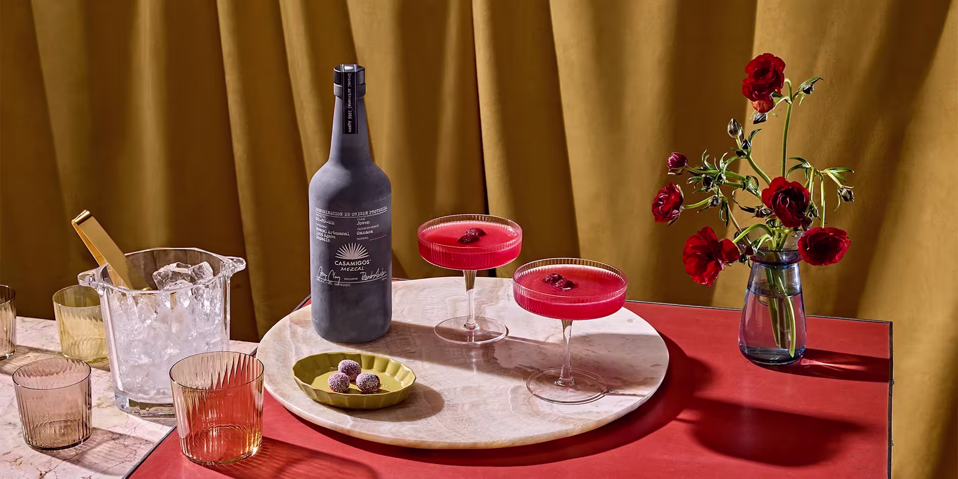 A marble tray with a dark bottle, two red cocktails in coupe glasses, a small plate with chocolates, and glassware, beside a vase with red flowers on a red surface.