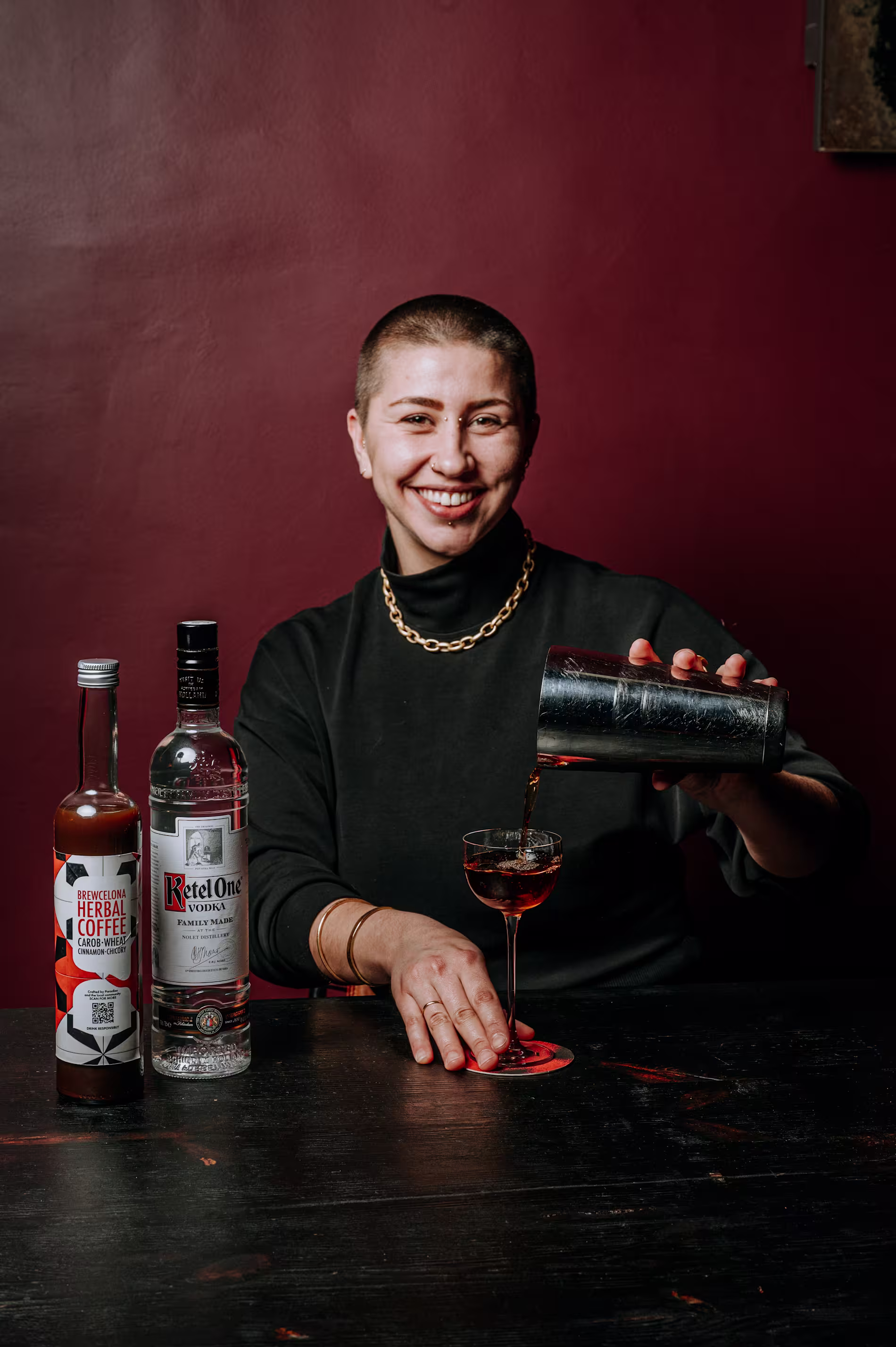 Persona sonriendo mientras sirve un cóctel desde una coctelera. Sobre la mesa, botellas de Ketel One y Brewcelona Herbal Coffee frente a una pared burdeos.