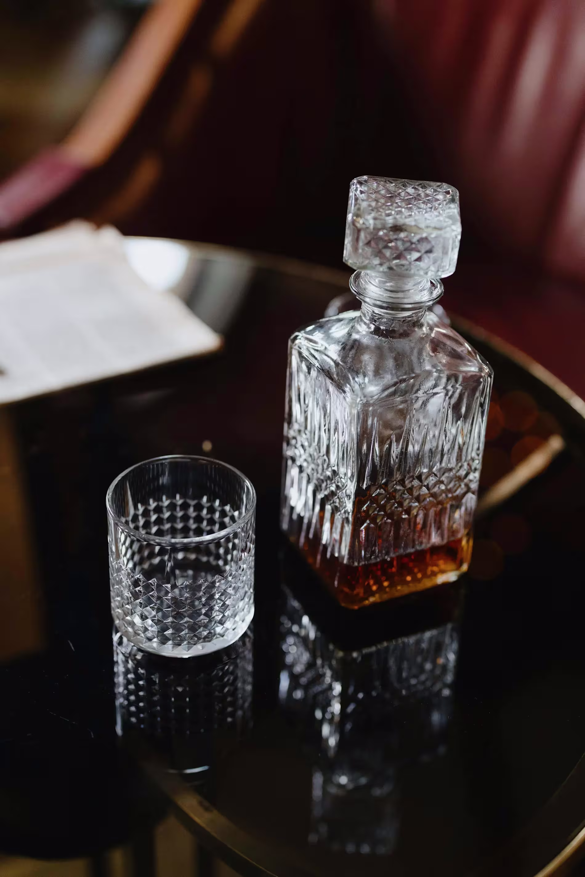 A whisky filled decanter sits next to an empty glass on a wooden table.