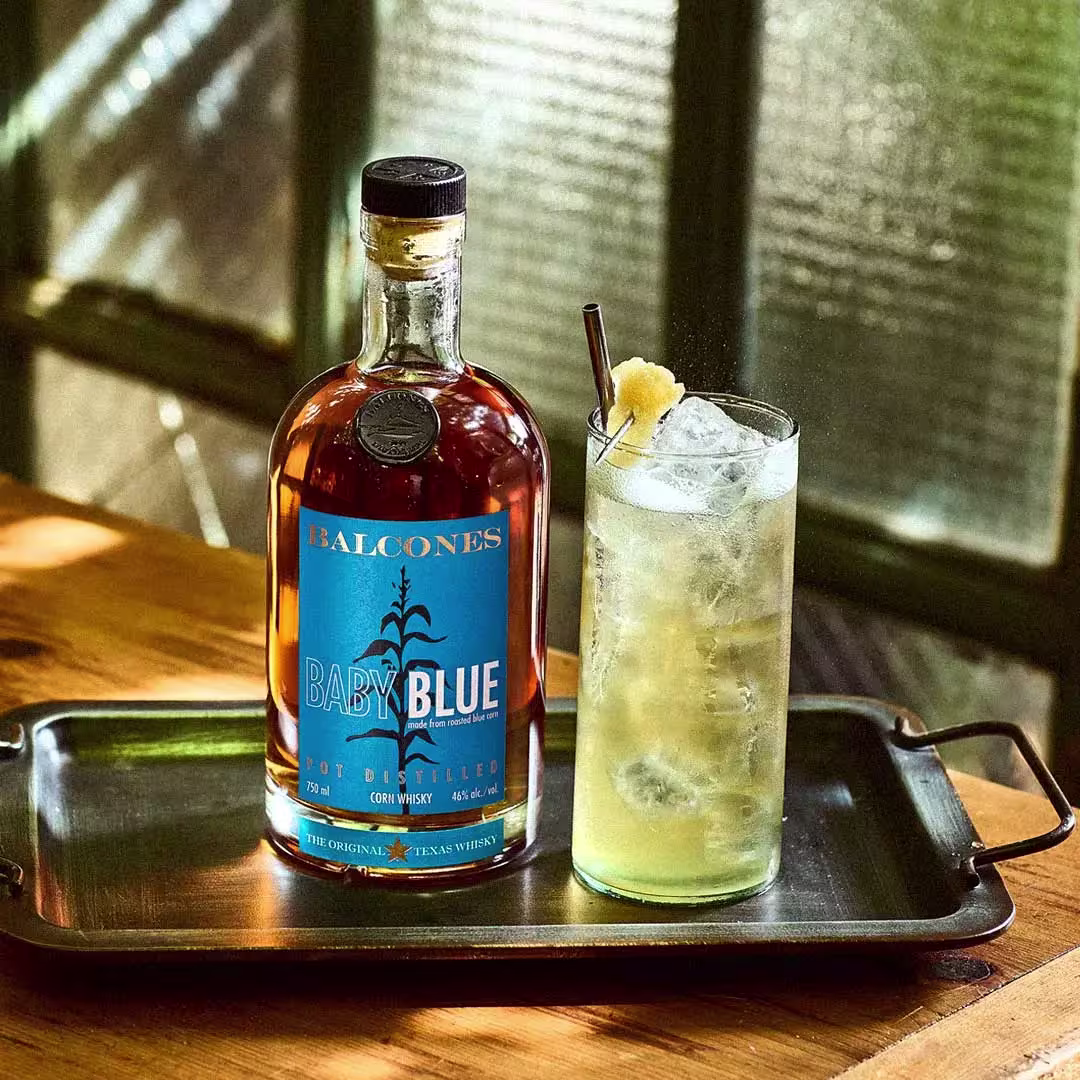 A bottle of Balcones Baby Blue whiskey and a tall glass of iced cocktail with a garnish on a metal tray, set on a wooden table by a window.