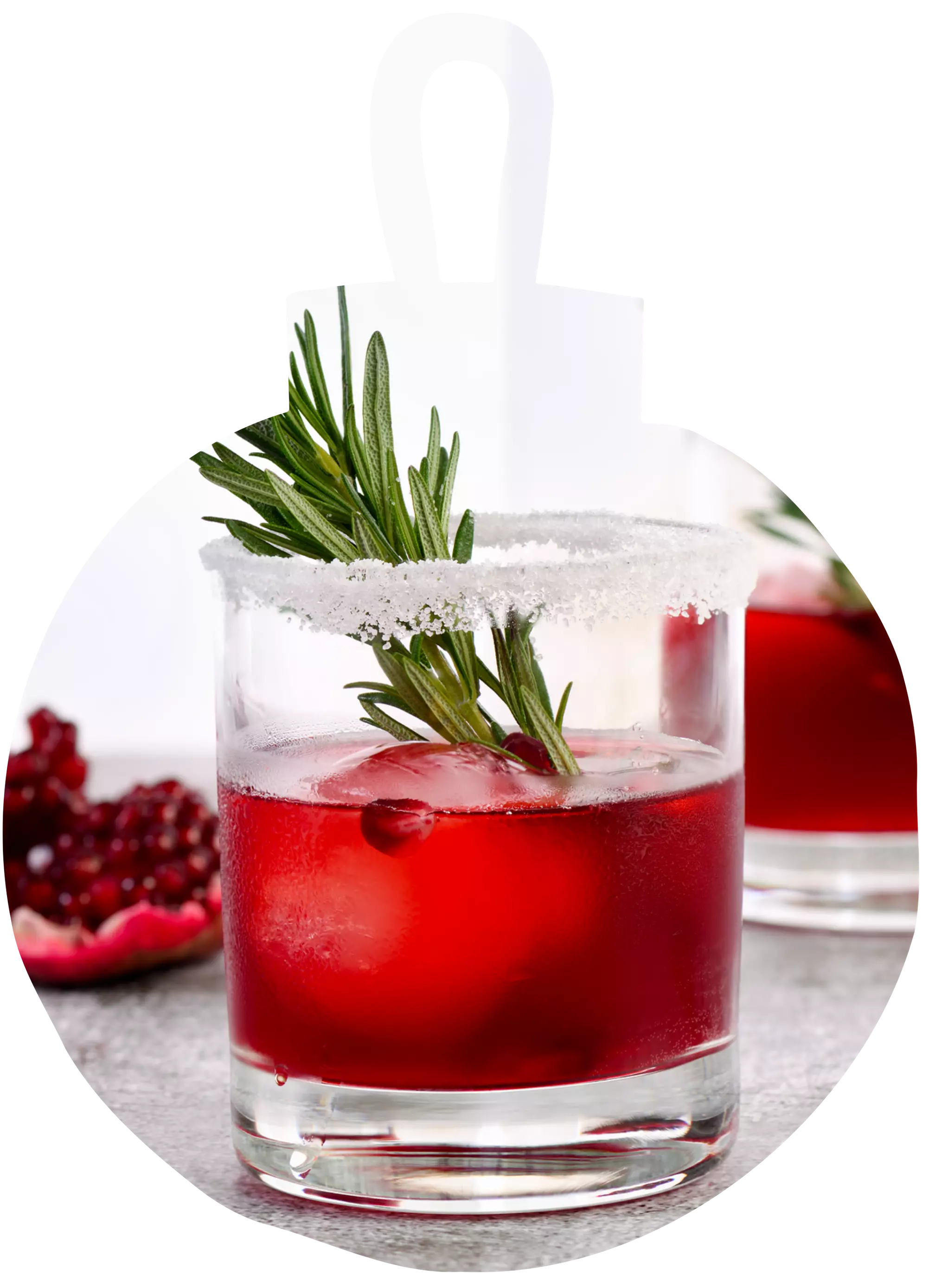 A glass of red cocktail with ice, garnished with a rosemary sprig and a sugared rim. Pomegranate seeds and another drink are visible in the background.
