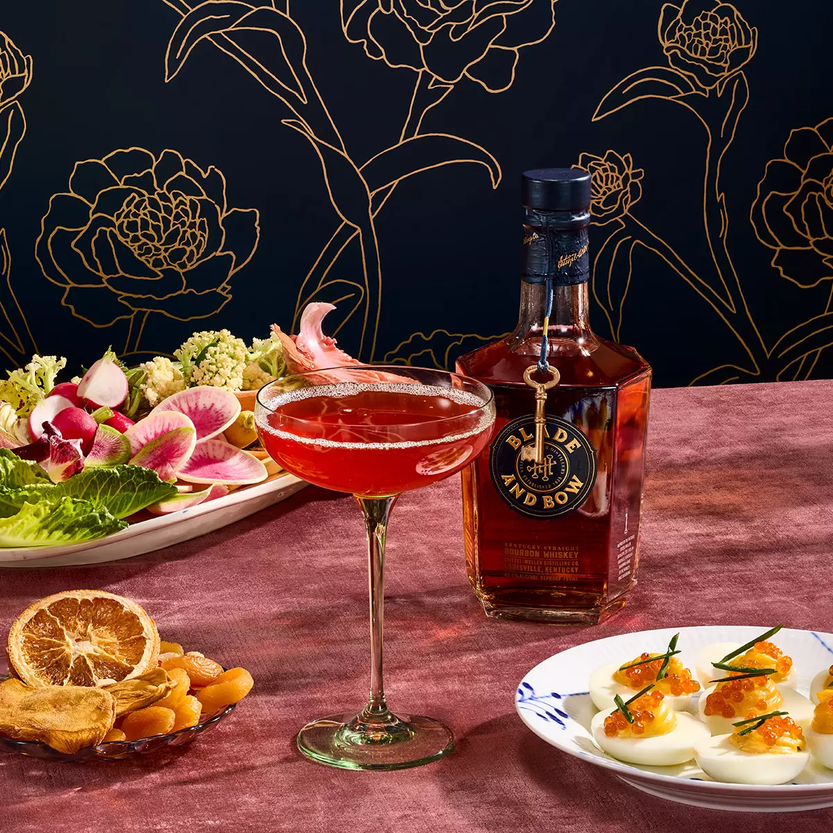 A cocktail in a coupe glass is placed on a table with a bottle of Blade and Bow whiskey, deviled eggs with garnish, dried fruits, and a plate of fresh vegetables.