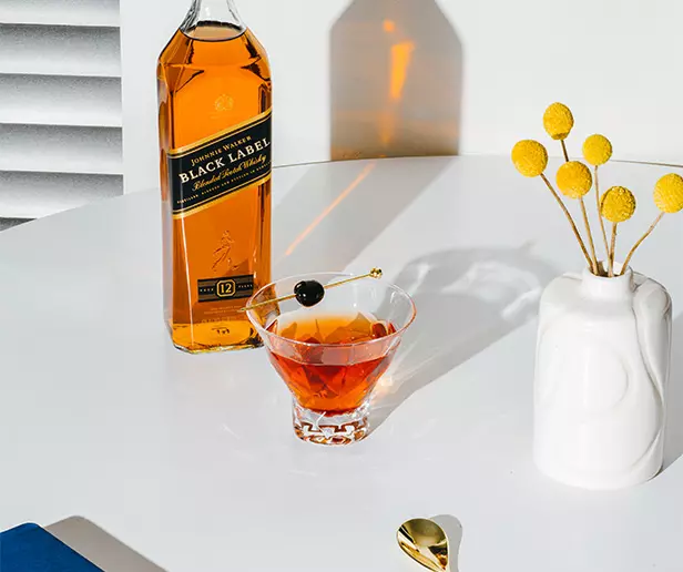 A bottle of Johnnie Walker Black Label whisky, a cocktail with a cherry garnish, a white vase with yellow billy balls, and a gold spoon on a white table.