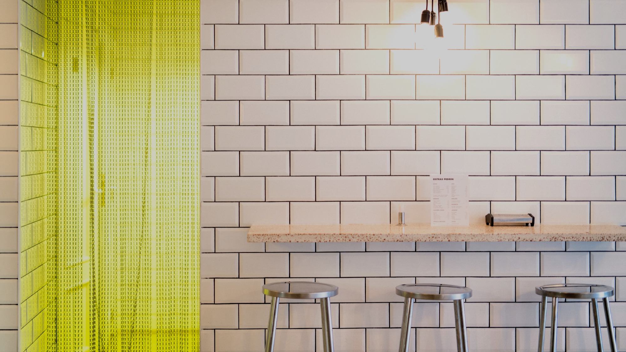Interior de Ostras Pedrín con barra alta, taburetes metálicos, azulejos blancos y cortina amarilla que aporta un toque moderno al espacio minimalista.