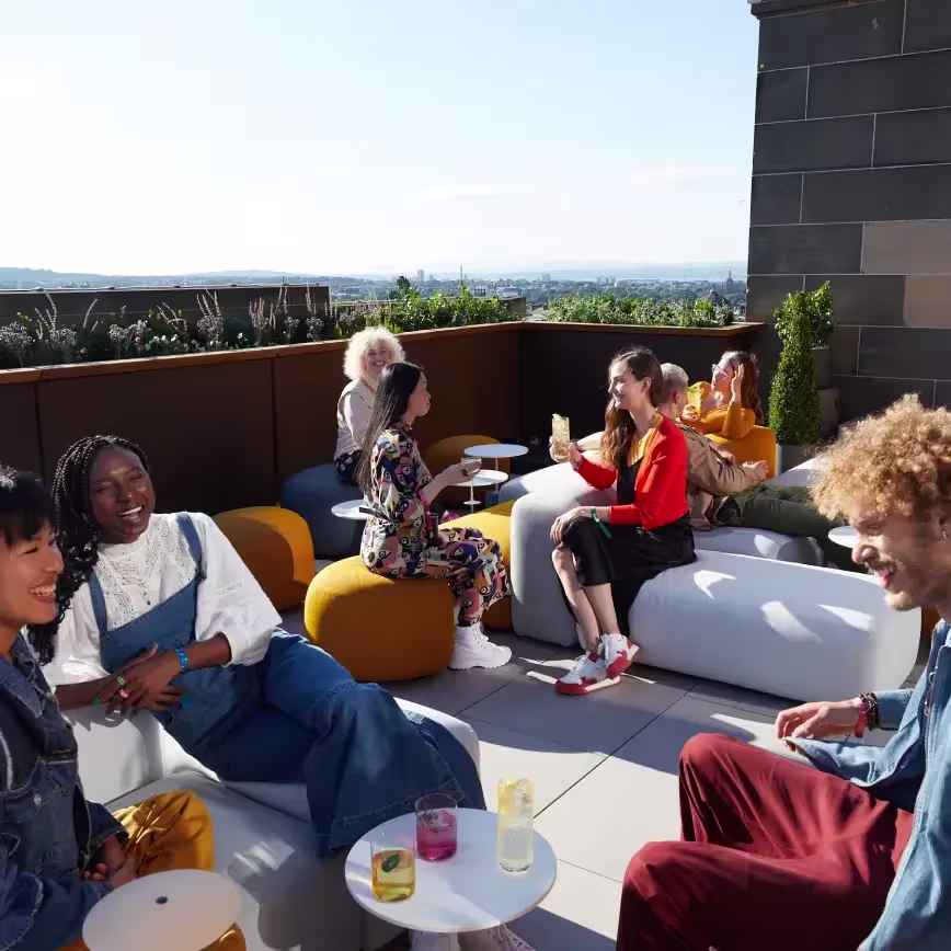 Un grupo de personas conversa en una azotea, tomando algo en un día soleado con un paisaje urbano de fondo.