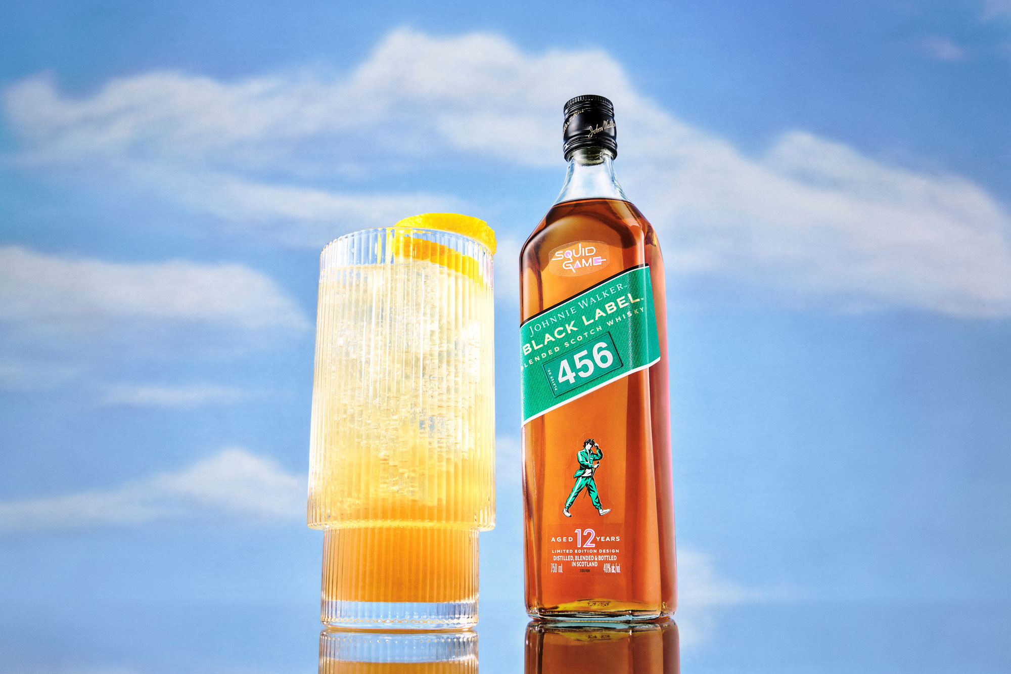 A bottle labeled “Black Label 456” stands next to a glass with a yellow cocktail and lemon garnish against a blue sky background.