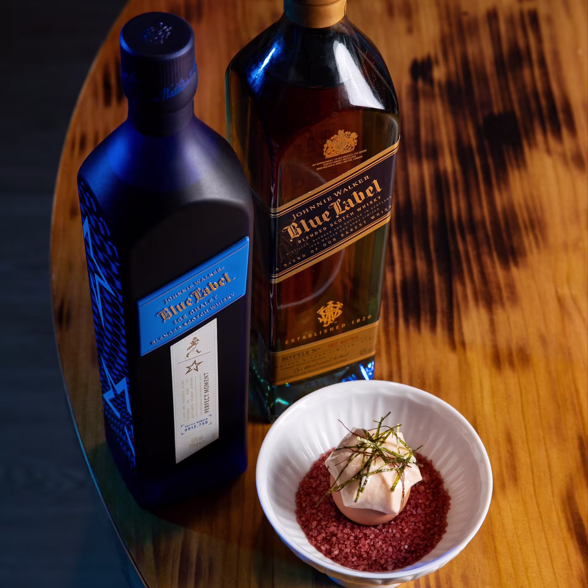 Dos botellas de whisky Johnnie Walker Blue Label acompañan un plato sofisticado, servido sobre sal rosada en un cuenco blanco, en una mesa de madera elegante.