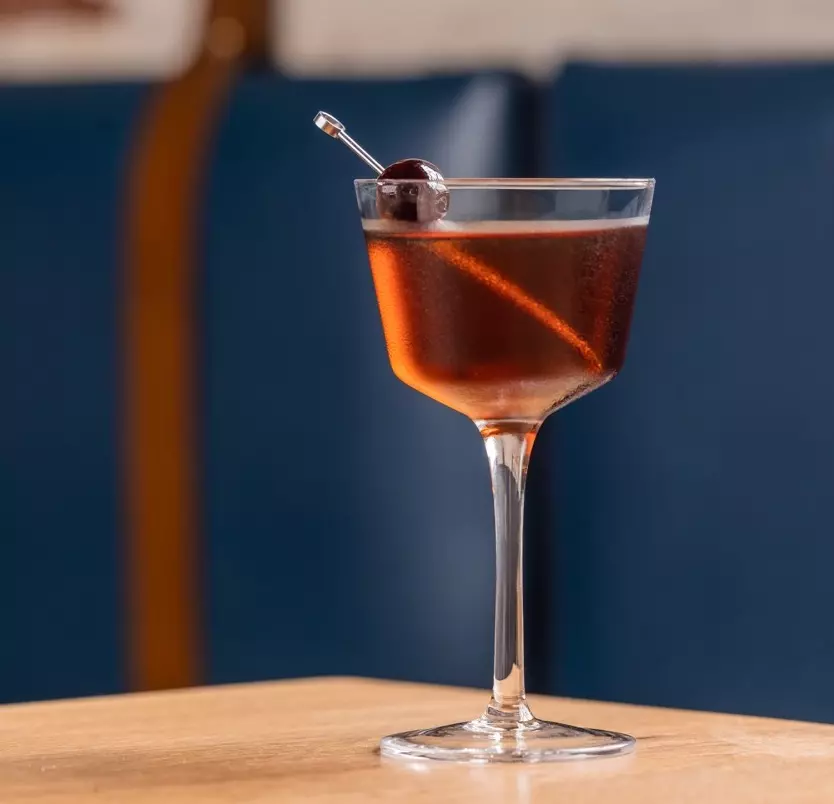 A cocktail in a stemmed glass with a dark liquid, garnished with a cherry on a pick and an orange peel, sits on a wooden table against a blue background.