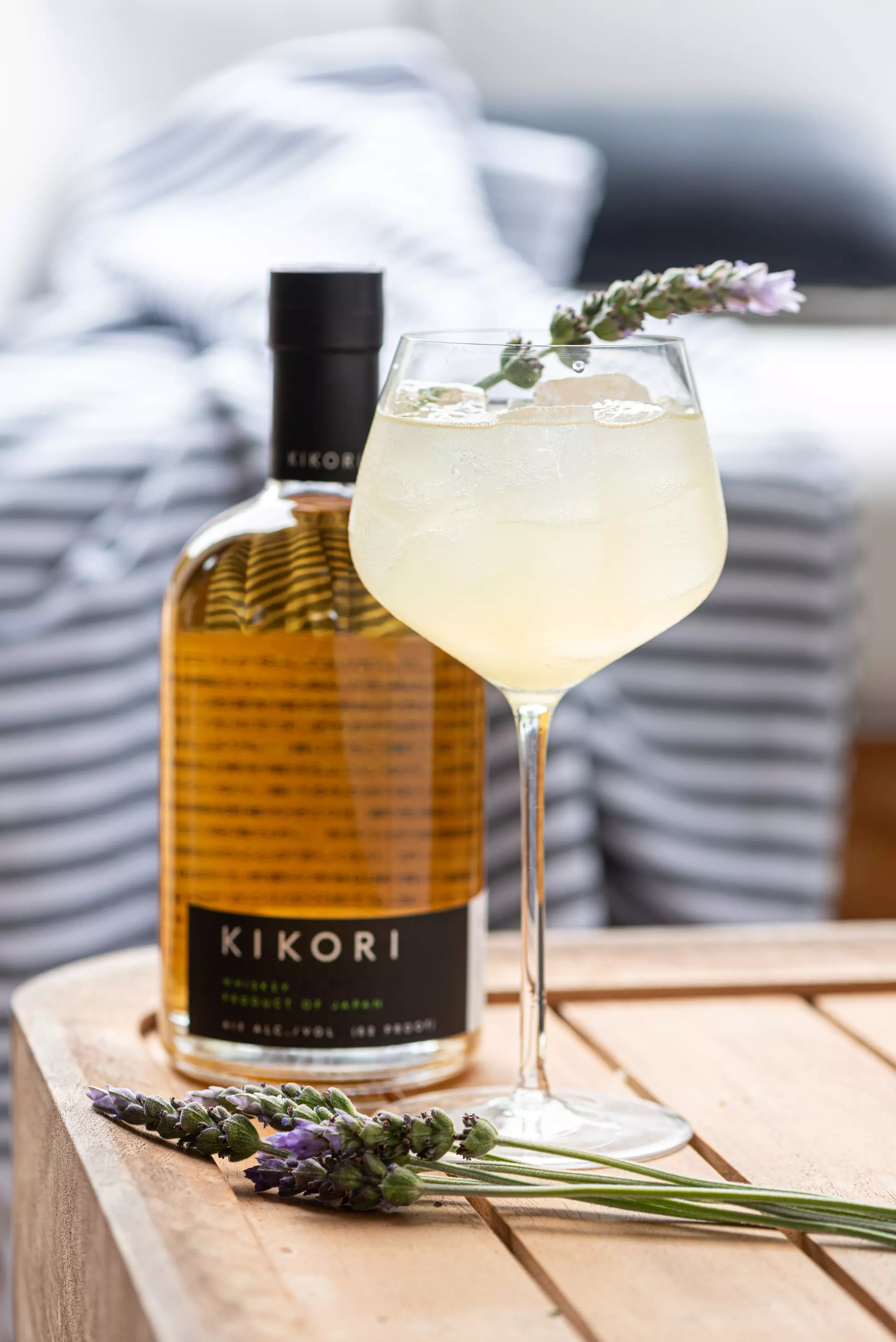 A glass of yellow cocktail with ice and a lavender garnish sits next to a bottle of Kikori whiskey on a wooden table.