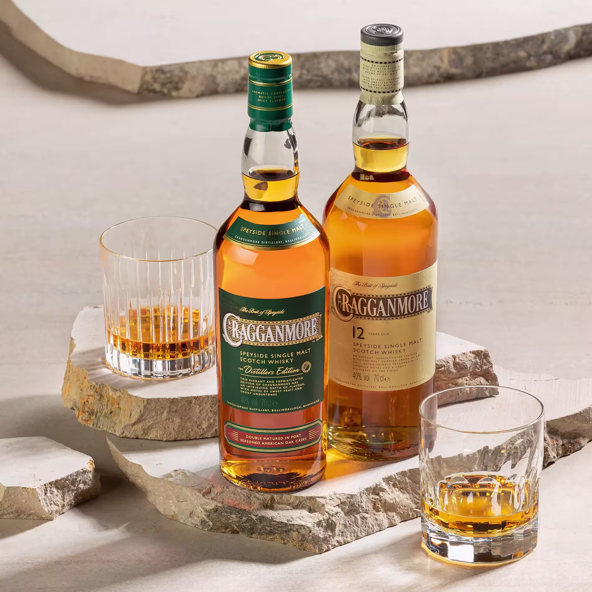Two bottles of Cragganmore Scotch whisky with three glasses containing whisky, arranged on flat stone slabs against a neutral background.