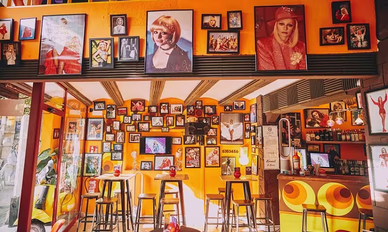 Interior de La Carra con paredes naranjas, fotos de iconos pop, mesas altas y una barra colorida, creando un ambiente vibrante y retro.