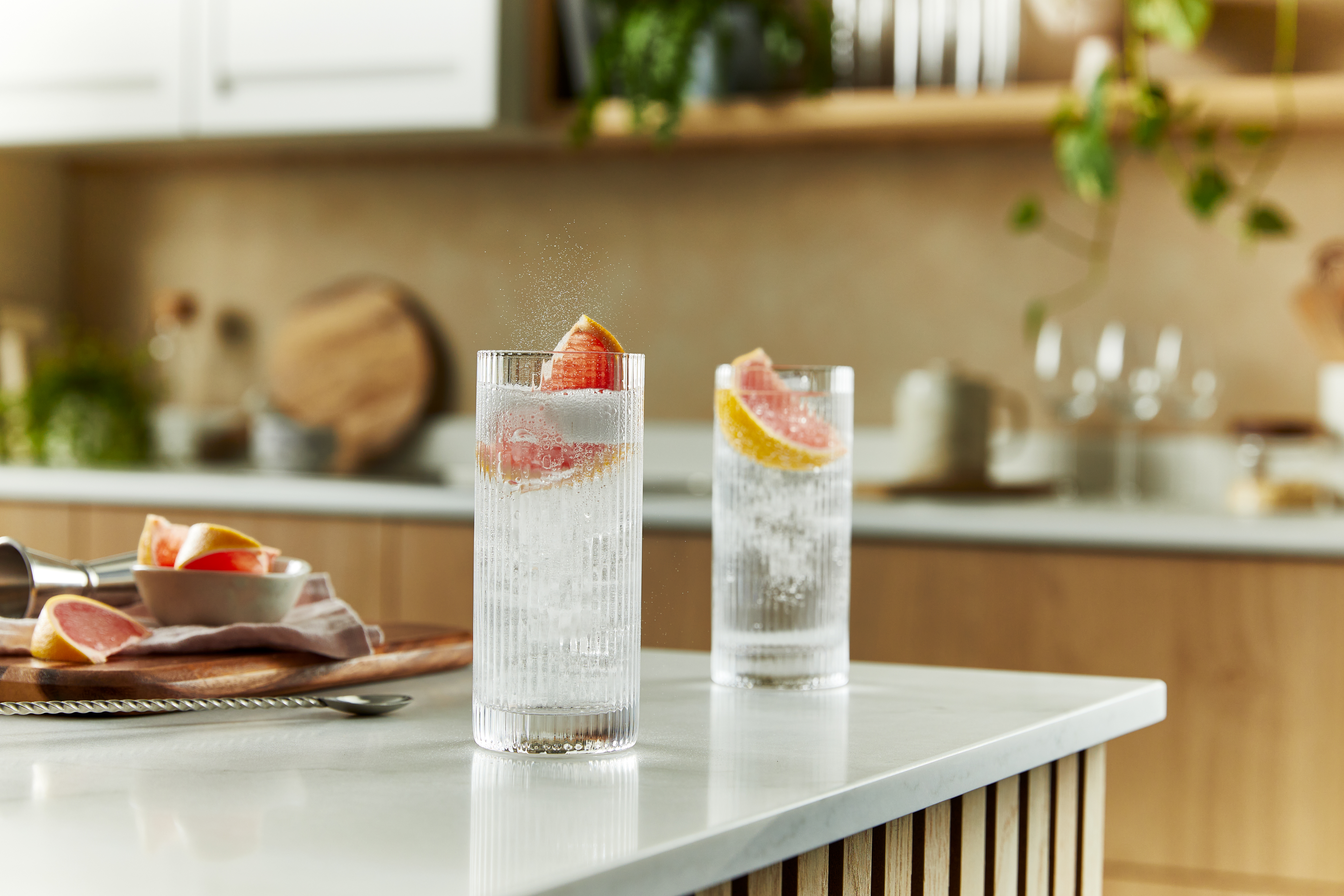 Two gin and tonic cocktails sit on a kitchen counter, in highball glasses with a wedge of pink grapefruit