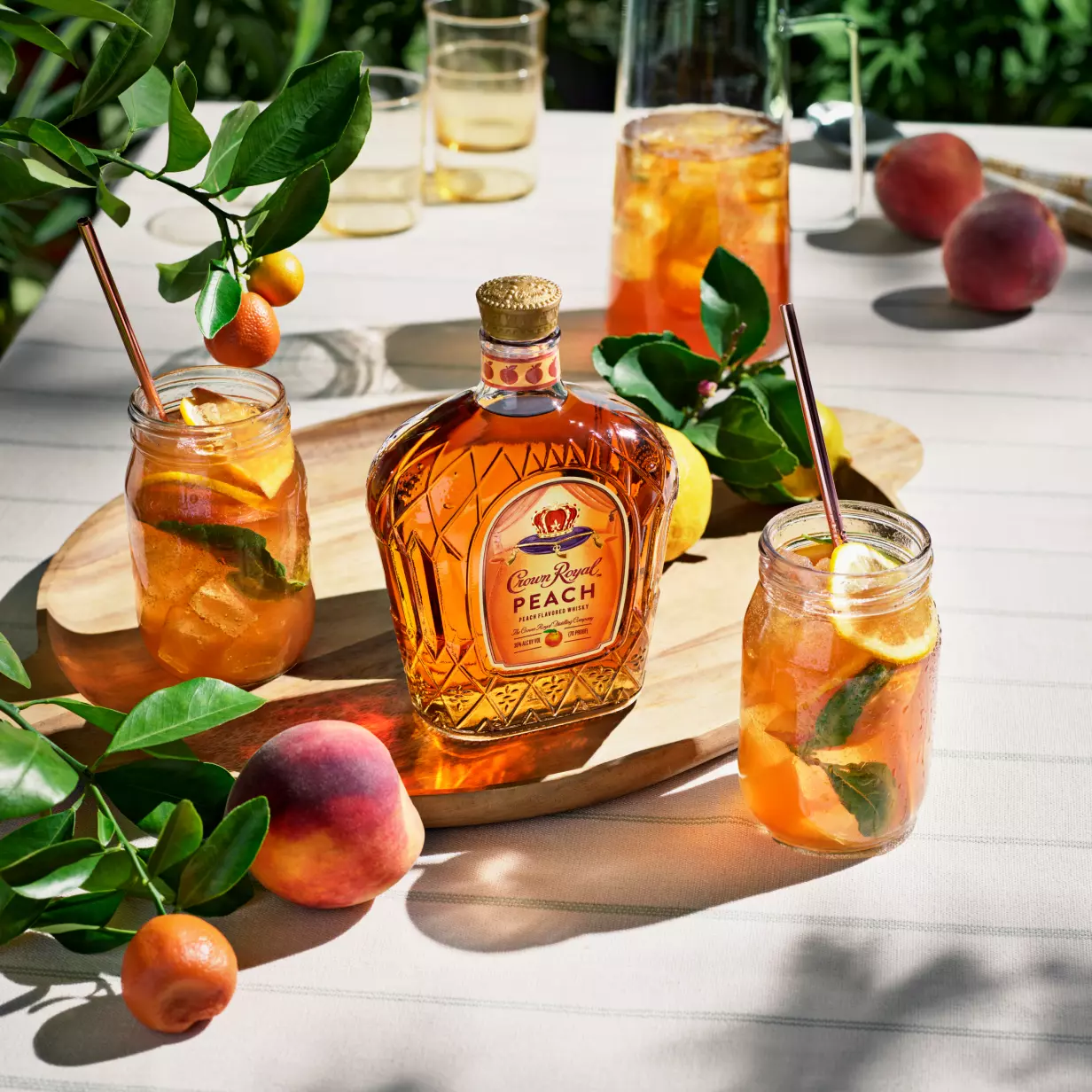 A bottle of Crown Royal Peach whiskey sits on a wooden board next to three mason jar cocktails with fruit and mint, surrounded by peaches and kumquats on a striped tablecloth.