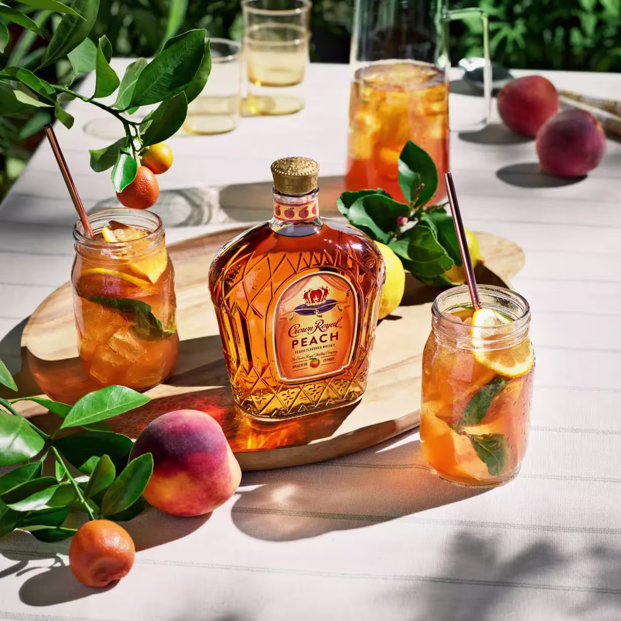 A bottle of Crown Royal Peach whiskey sits on a wooden board next to three mason jar cocktails with fruit and mint, surrounded by peaches and kumquats on a striped tablecloth.
