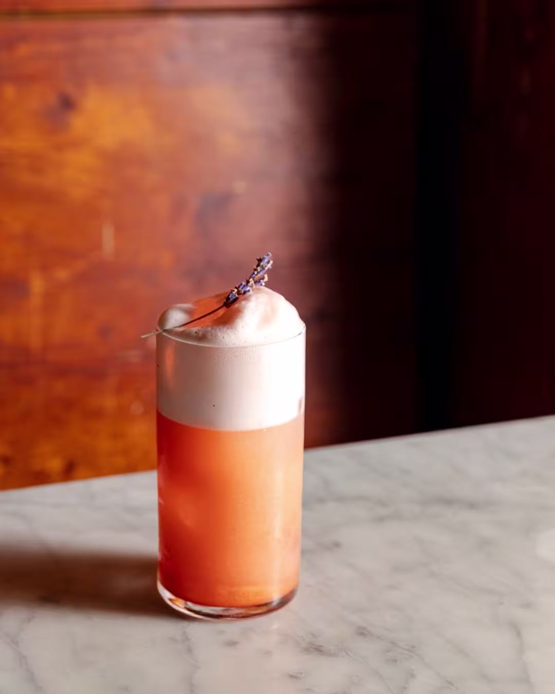 Cóctel anaranjado con espuma blanca, decorado con ramita de lavanda, servido en vaso alto sobre mesa de mármol con fondo de madera.