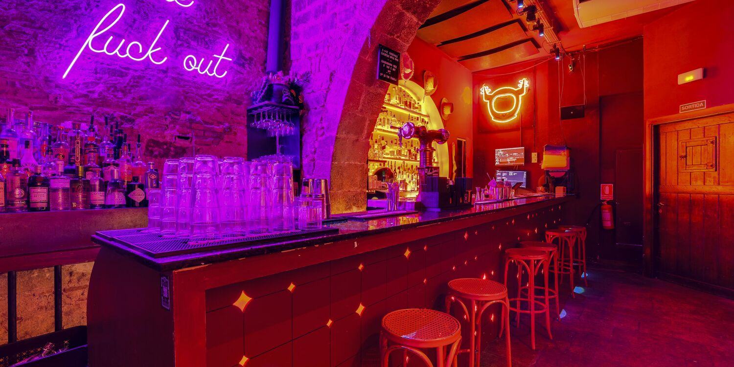 La imagen muestra un bar de ambiente vibrante, con luces de neón en tonos morados y rojos. La barra está llena de botellas y hay taburetes metálicos junto a ella. La decoración tiene un estilo urbano y moderno.