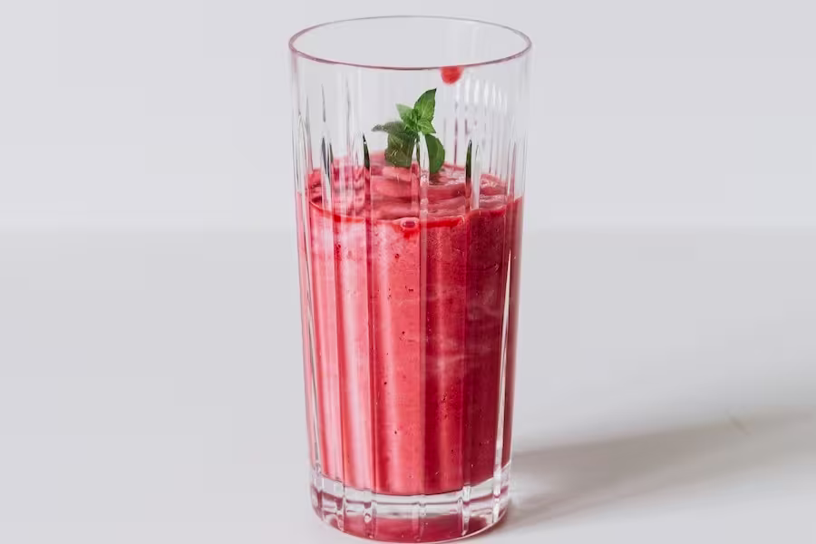 A tall, clear glass filled with a thick red smoothie, garnished with a small sprig of mint, on a white surface.