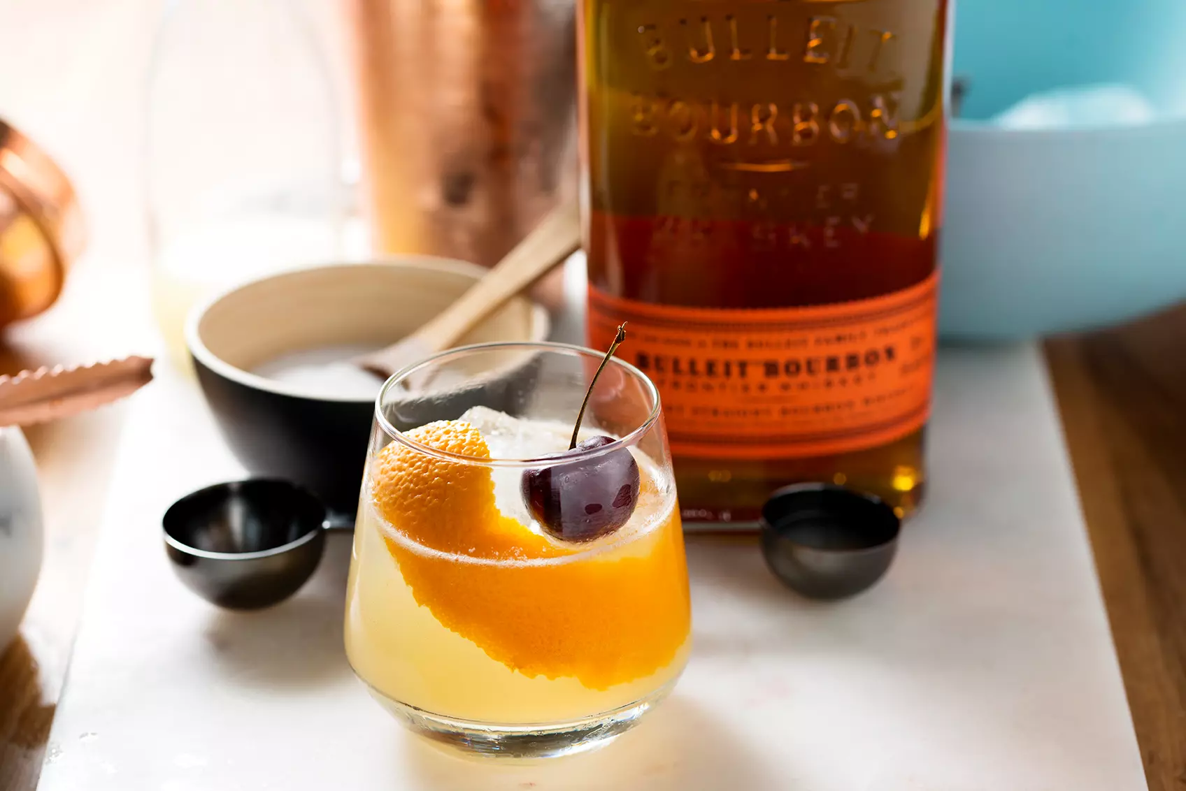 A glass of whiskey cocktail garnished with an orange peel and cherry sits on a table next to a bottle of Bulleit Bourbon and measuring cups.