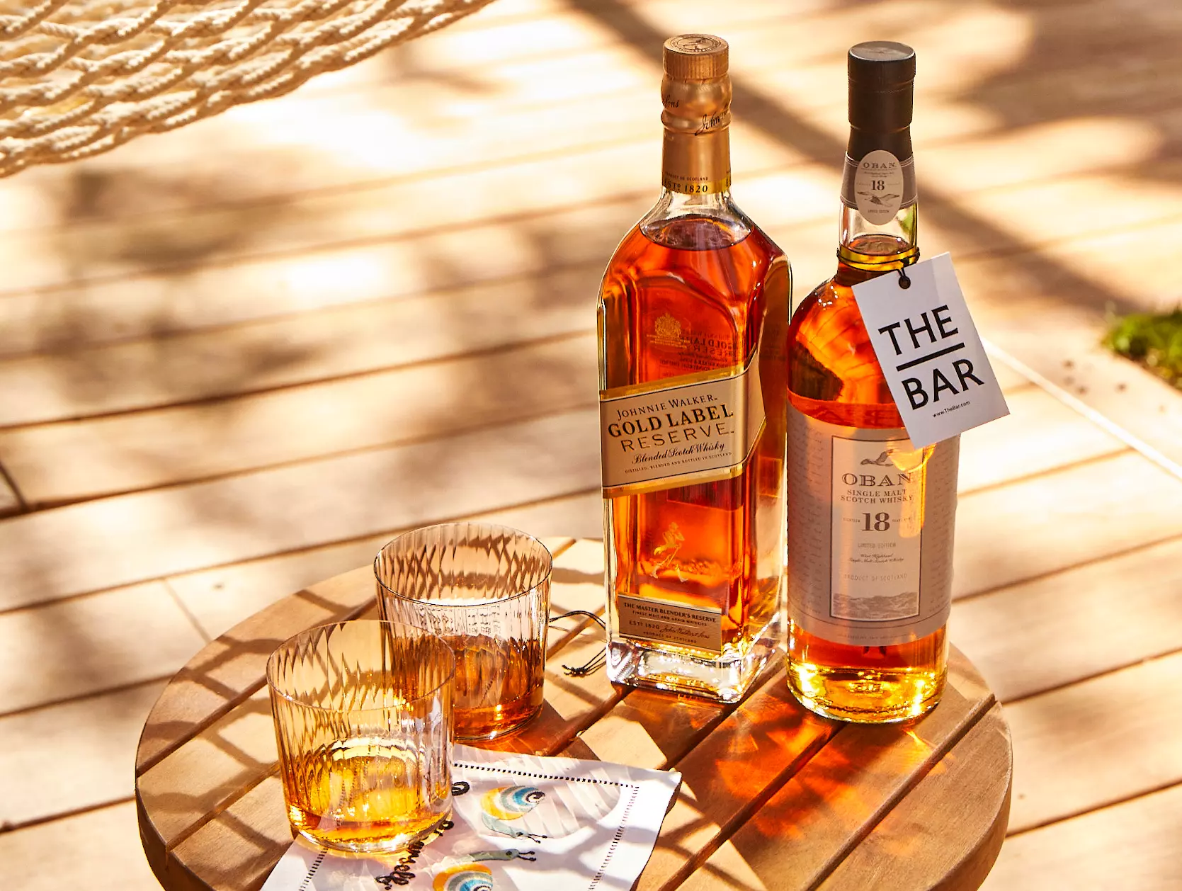 Two bottles of whisky and two filled glasses sit on a round wooden table in sunlight. A napkin is also on the table.