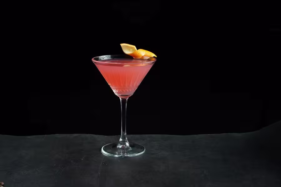 A pink cocktail served in a martini glass, garnished with two orange slices, set against a dark background.