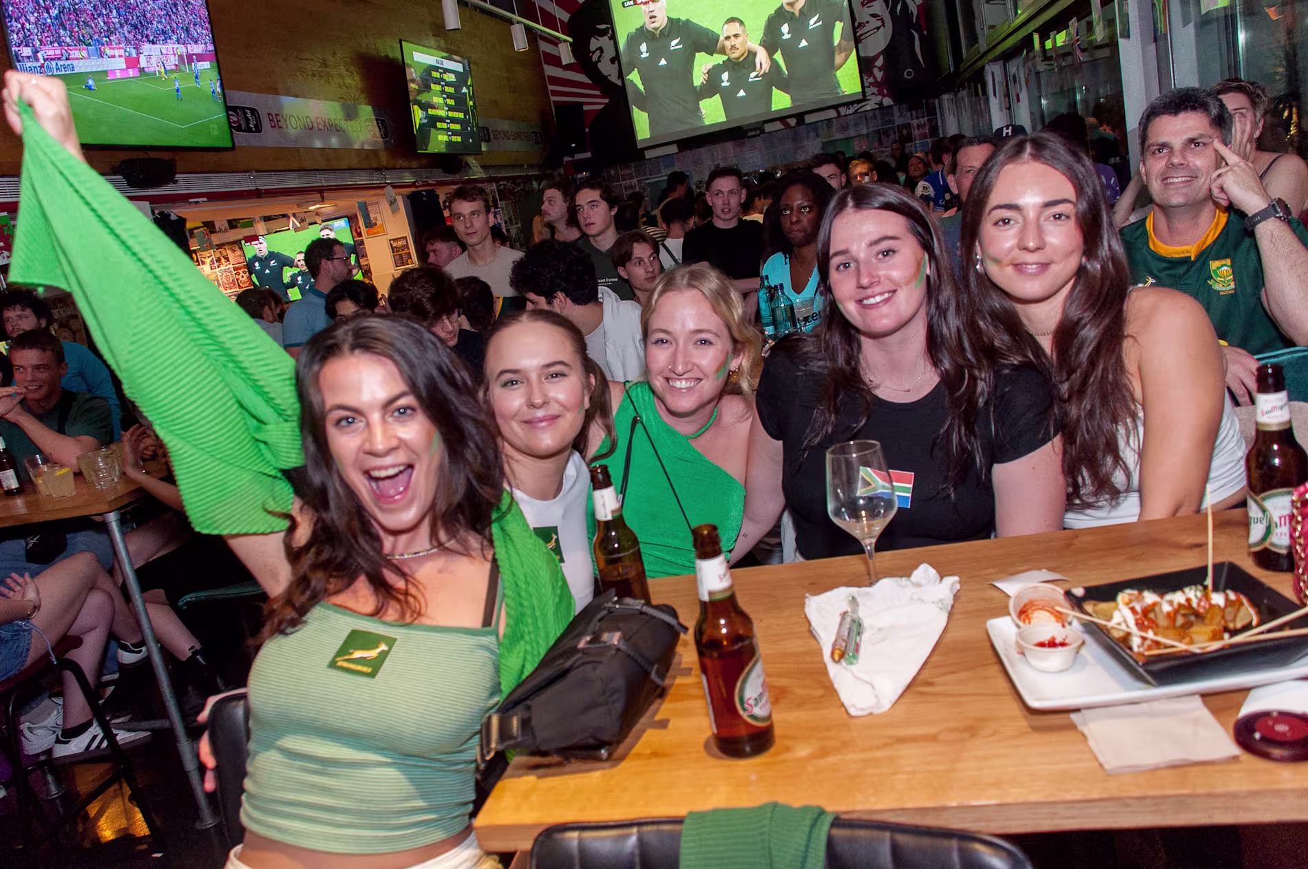 La imagen muestra un grupo de personas en el interior del bar belushis disfrutando de un evento deportivo, hay varias bebidas y alimentos en las mesas, lo que indica una reunión social."