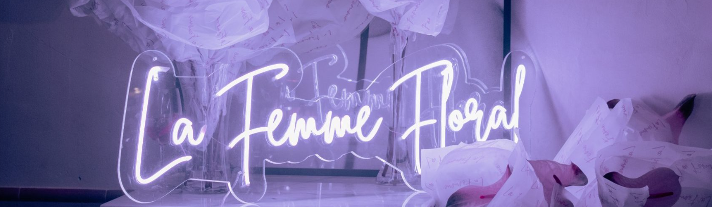 La imagen muestra un cartel luminoso con el nombre "La Femme Floral" sobre un fondo decorado con flores blancas. El diseño tiene un tono romántico y elegante, con una atmósfera acogedora.